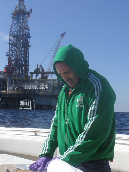 The research team's second sampling expedition in December 2012, was more challenging due to the close proximity of a BP survey vessel, Olympic Triton, which was operating two ROVs, and a mobile offshore deep drilling unit. The rig and vessel each require a reasonable stand-off distance for safety precautions. WHOI's Chris Reddy is shown here, with the drilling rig behind him. (Photo by Bob Nelson, Woods Hole Oceanographic Institution)